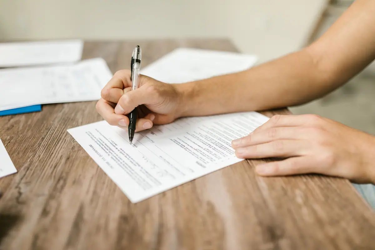 A hand filling in a paper form with a pen on a wooden desk