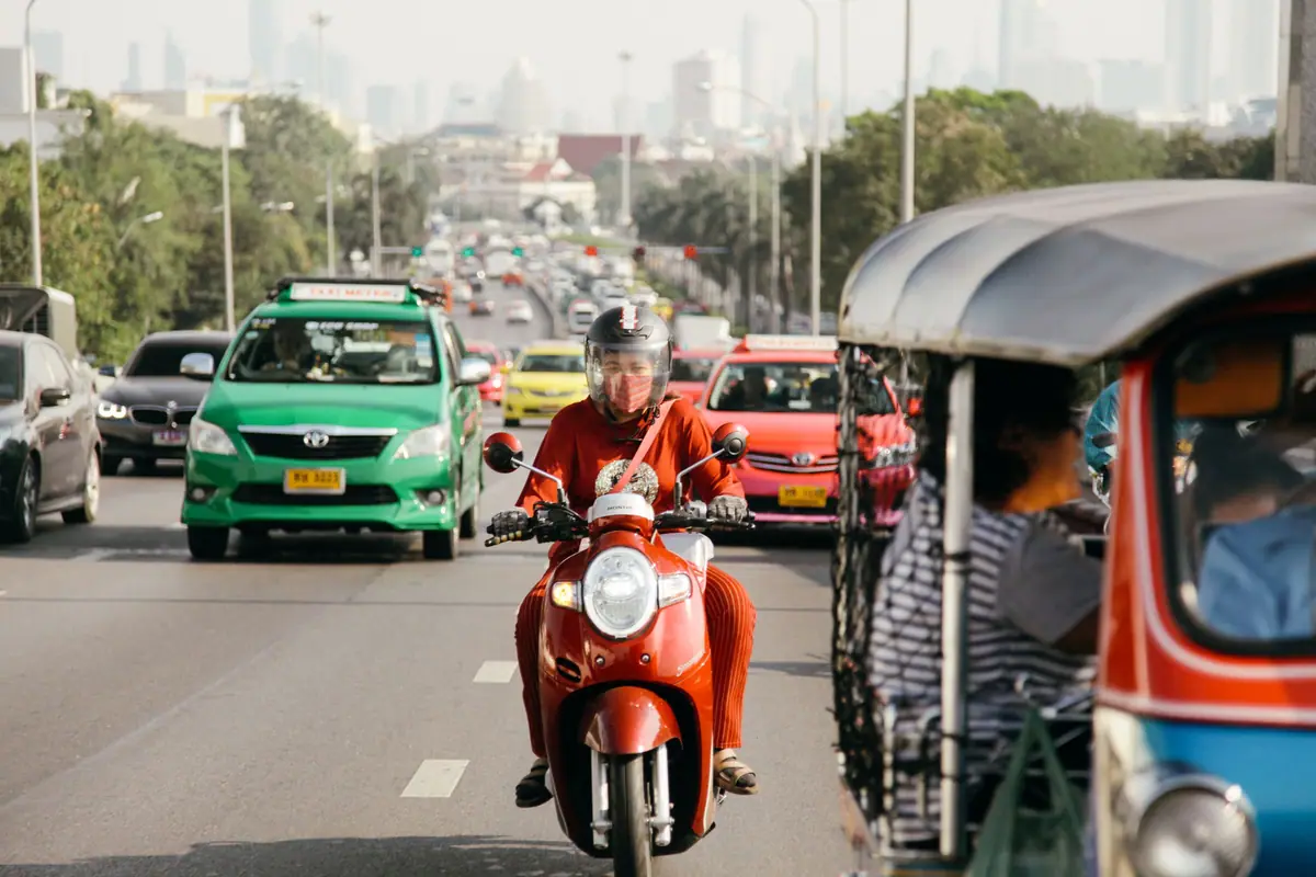 A rider on a red scooter in Bangkok traffic next to a tuk-tuk
